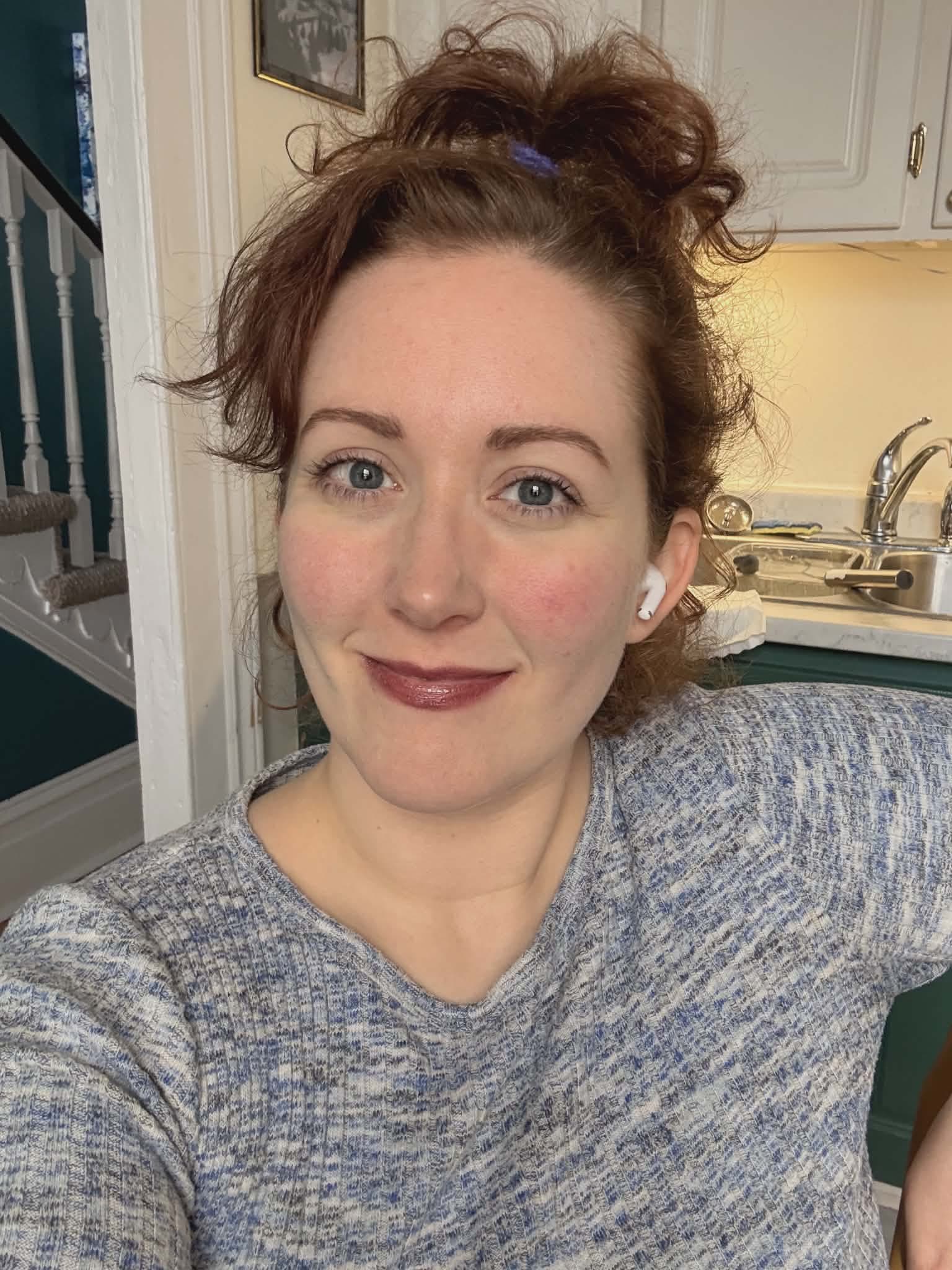 A woman with curly brown hair tied up smiles at the camera indoors. She wears a gray long-sleeve shirt and a white wireless earbud. A kitchen with a sink and cabinets is visible behind her, along with a staircase in the background.
