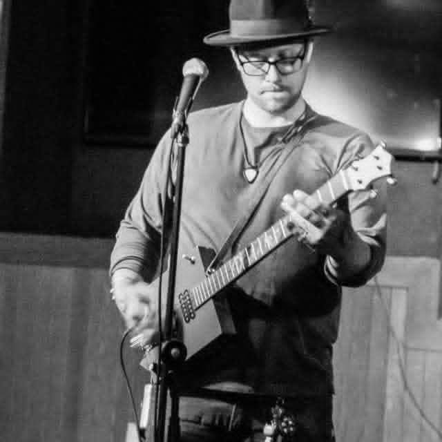 Black-and-white photo of a man wearing a hat and glasses, playing an electric guitar. He stands near a microphone on a stage, looking down at the instrument, with a blurred indoor background behind him.