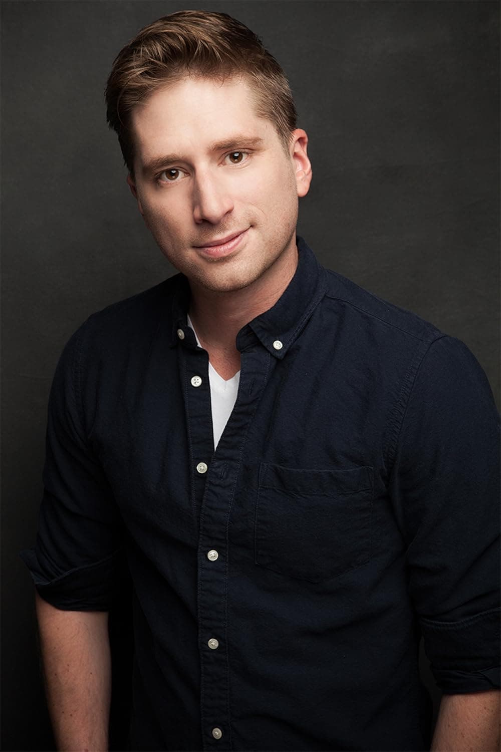 A studio portrait of a person with short light brown hair, looking at the camera with a slight smile. They wear a dark navy button-up shirt over a white V-neck T-shirt. The background is a solid dark gray, and the image is evenly lit with soft studio lighting.