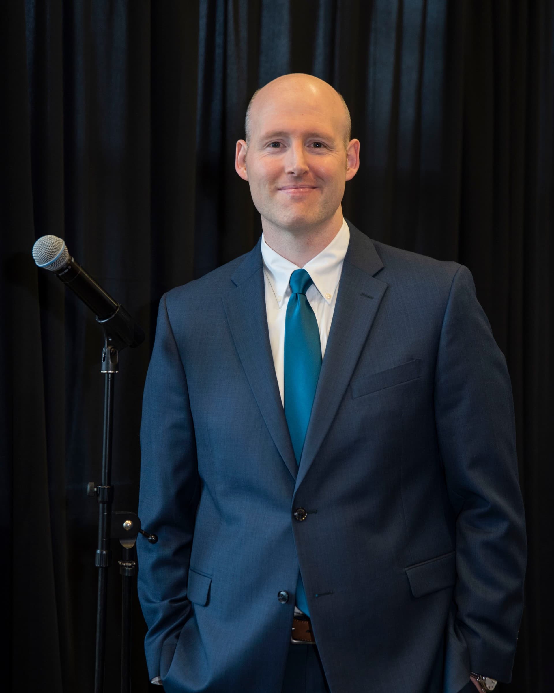 A bald man in a blue suit and teal tie stands beside a microphone on a stand. He faces the camera with a slight smile, with a black curtain backdrop behind him.