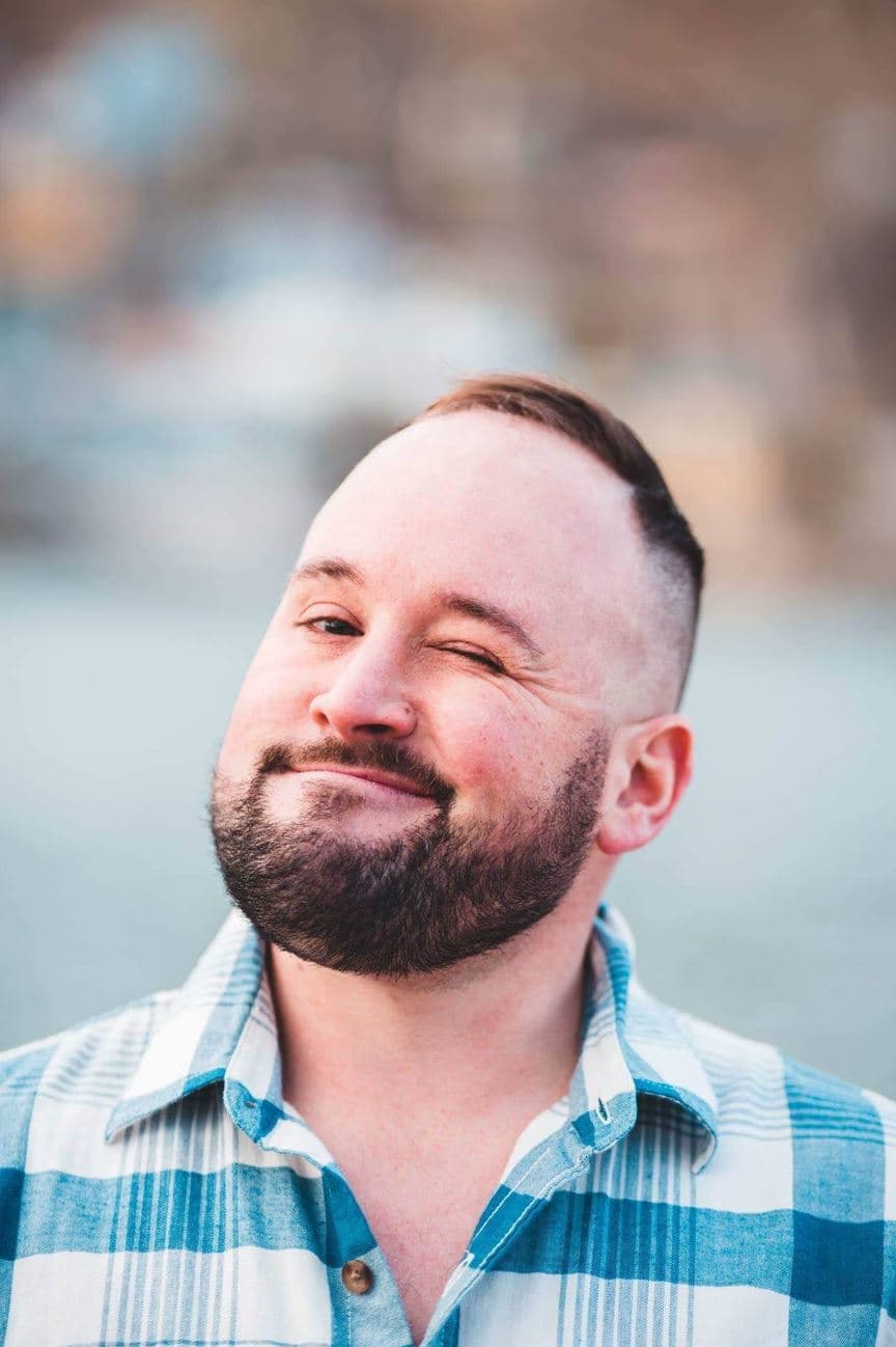 A man with a short haircut and full beard smiles while winking at the camera. He wears a blue and white plaid shirt, with a softly blurred outdoor background behind him.