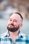 A man with a short haircut and full beard smiles while winking at the camera. He wears a blue and white plaid shirt, with a softly blurred outdoor background behind him.