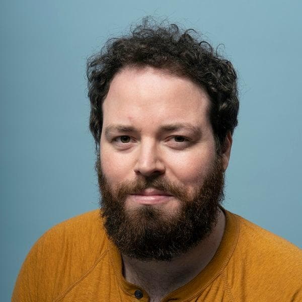 A head-and-shoulders portrait of a person with curly dark hair and a full dark beard, looking at the camera with a slight closed-mouth smile. They wear a mustard-yellow henley shirt. The background is a plain light blue backdrop with even studio lighting.