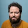 A head-and-shoulders portrait of a person with curly dark hair and a full dark beard, looking at the camera with a slight closed-mouth smile. They wear a mustard-yellow henley shirt. The background is a plain light blue backdrop with even studio lighting.