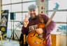 A man wearing a gray flat cap and maroon sweatshirt holds a hollow-body electric guitar and smiles while making a peace sign with his hand. He stands indoors near large garage-style windows, with a speaker and a sign visible in the background.