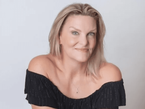 Studio portrait of a person with shoulder-length blonde hair and blue eyes, wearing an off-the-shoulder black top and a small pendant necklace. They face the camera with a slight smile against a plain light gray background.