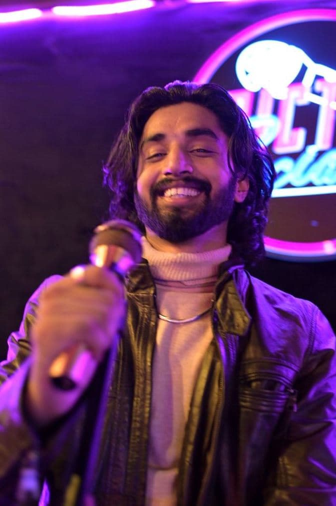 A person with shoulder-length dark hair and a beard smiles while holding a microphone toward the camera. They wear a leather jacket over a light turtleneck shirt. Purple and pink neon lights and a blurred sign glow in the background.