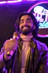 A person with shoulder-length dark hair and a beard smiles while holding a microphone toward the camera. They wear a leather jacket over a light turtleneck shirt. Purple and pink neon lights and a blurred sign glow in the background.