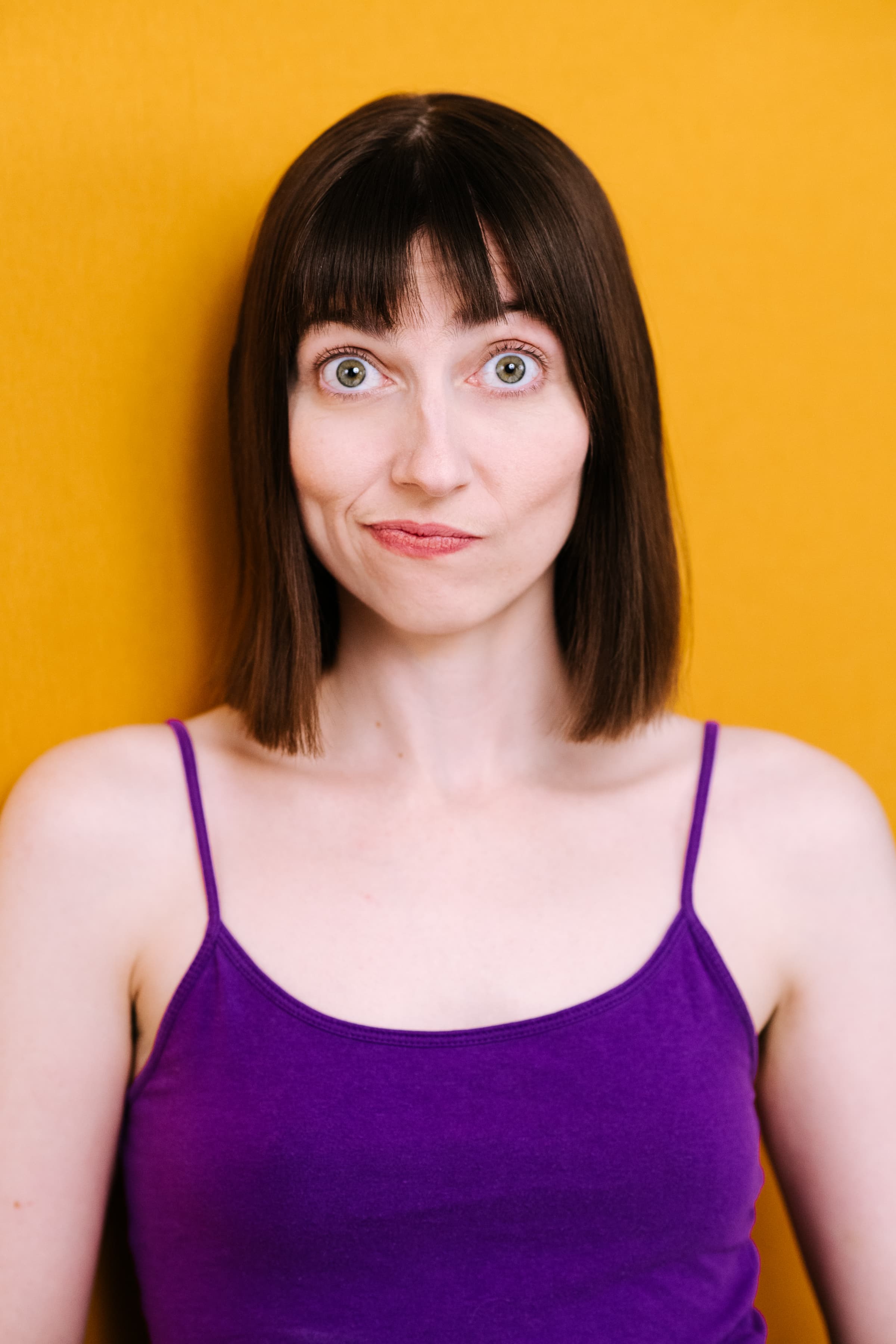 A person with straight, shoulder-length dark brown hair and bangs faces forward against a solid yellow background, wearing a purple tank top. Their eyes are wide open, and their lips are slightly pursed.