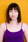 A person with straight, shoulder-length dark brown hair and bangs faces forward against a solid yellow background, wearing a purple tank top. Their eyes are wide open, and their lips are slightly pursed.
