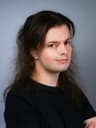 A person with long dark hair faces slightly to the side with arms crossed, wearing a black T-shirt. They are photographed against a plain gray background with a neutral expression.