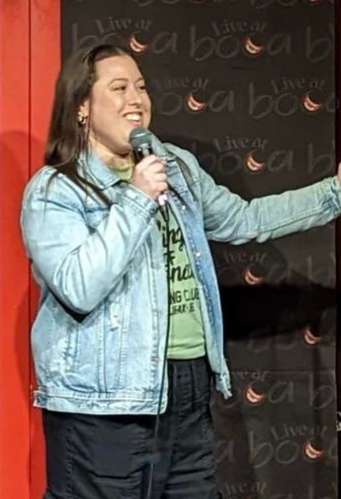 A person stands on stage holding a microphone, speaking with one arm extended. They wear a denim jacket over a green shirt and black pants. A backdrop with repeated “Live at Boabab” text and crescent moon logos is behind them.