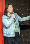 A person stands on stage holding a microphone, speaking with one arm extended. They wear a denim jacket over a green shirt and black pants. A backdrop with repeated “Live at Boabab” text and crescent moon logos is behind them.