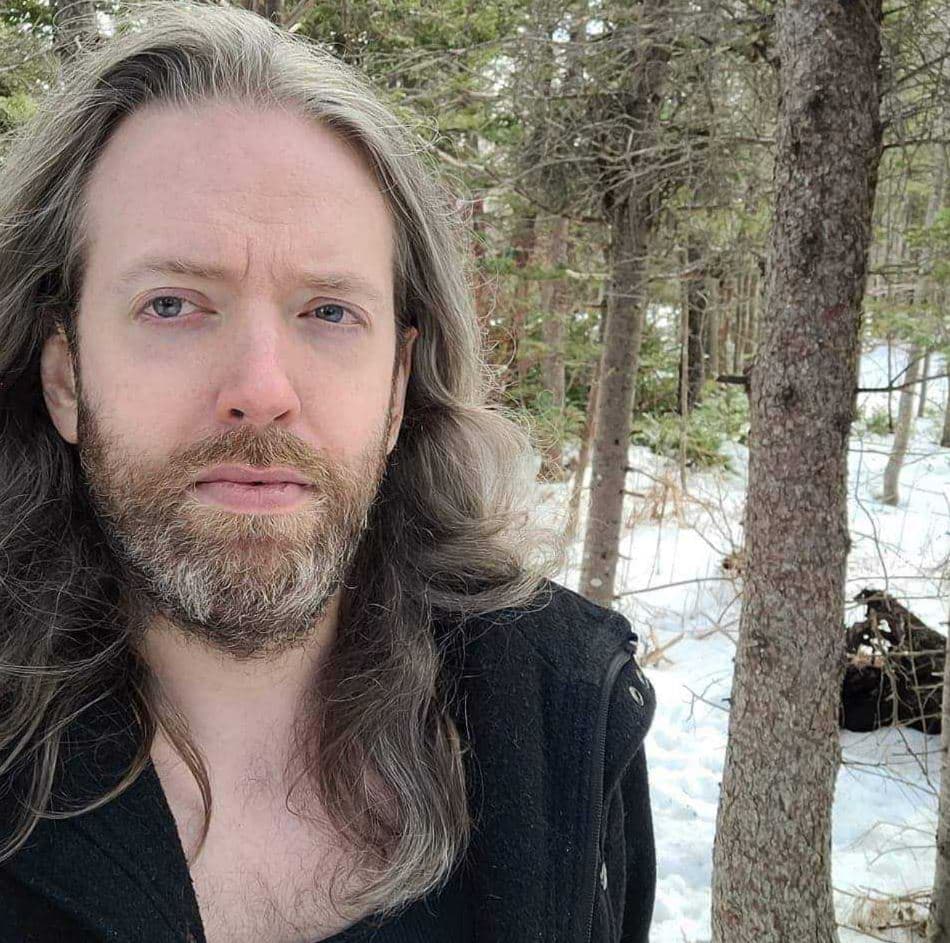 A person with long light-brown hair and a beard looks at the camera outdoors. They wear a dark jacket, standing in a snowy forest with trees and patches of snow visible in the background.