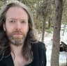 A person with long light-brown hair and a beard looks at the camera outdoors. They wear a dark jacket, standing in a snowy forest with trees and patches of snow visible in the background.