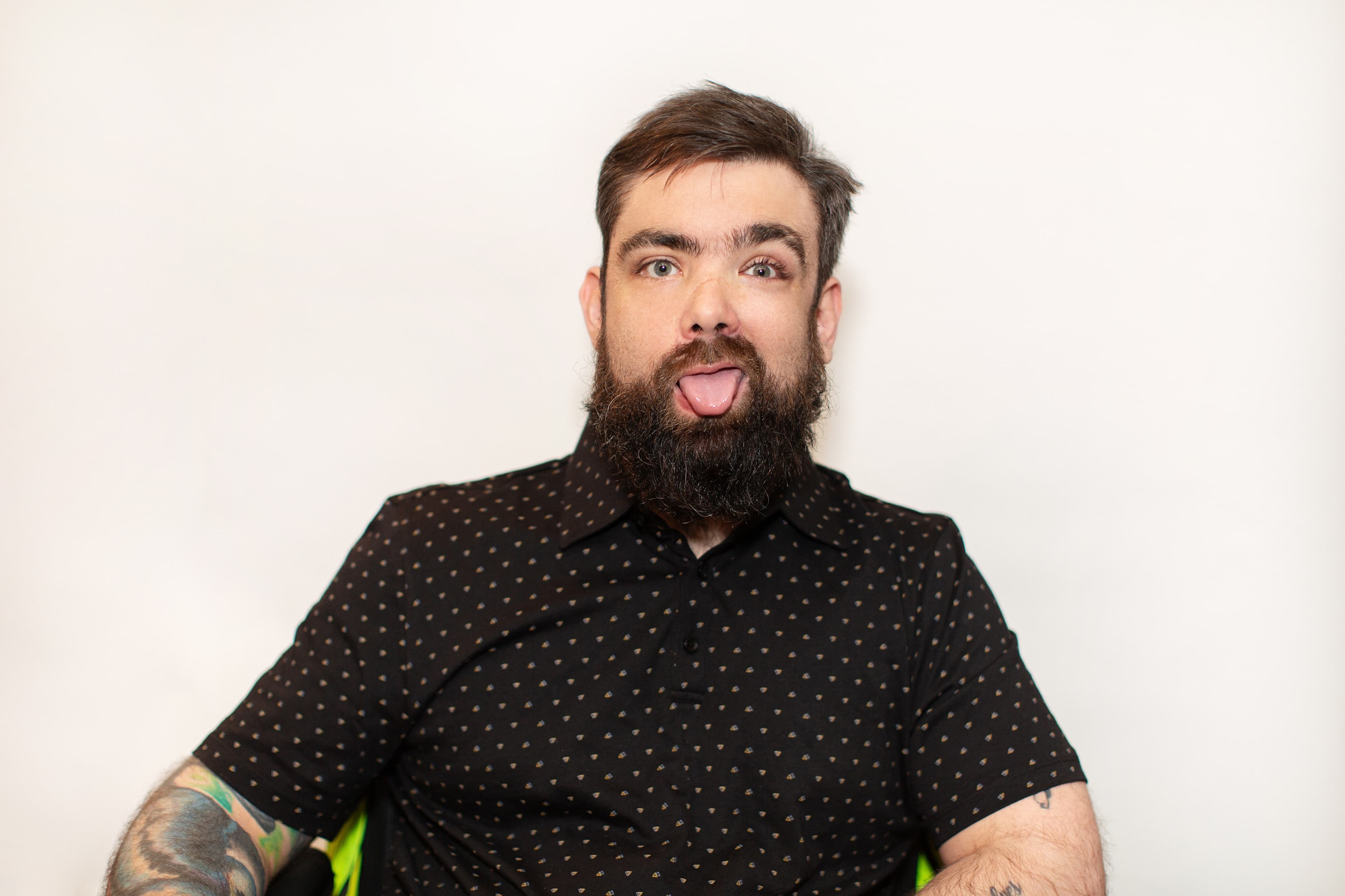 A man with short brown hair and a full beard faces the camera, sticking out his tongue. He wears a black short-sleeve button-up shirt with a small dotted pattern. Tattoos are visible on both forearms. The background is plain and light-colored.