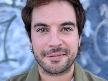 A close-up head-and-shoulders portrait of a person with short dark hair, light-colored eyes, and short facial hair, looking at the camera with a slight smile. They wear an olive-green jacket. The background is an out-of-focus blue wall with graffiti-like markings.