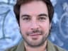 A close-up head-and-shoulders portrait of a person with short dark hair, light-colored eyes, and short facial hair, looking at the camera with a slight smile. They wear an olive-green jacket. The background is an out-of-focus blue wall with graffiti-like markings.