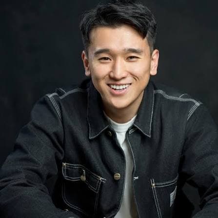 Studio headshot of a smiling person with short dark hair, wearing a dark denim-style jacket over a light shirt. The person is leaning slightly forward with arms resting in front, against a plain dark background.