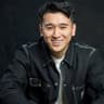 Studio headshot of a smiling person with short dark hair, wearing a dark denim-style jacket over a light shirt. The person is leaning slightly forward with arms resting in front, against a plain dark background.