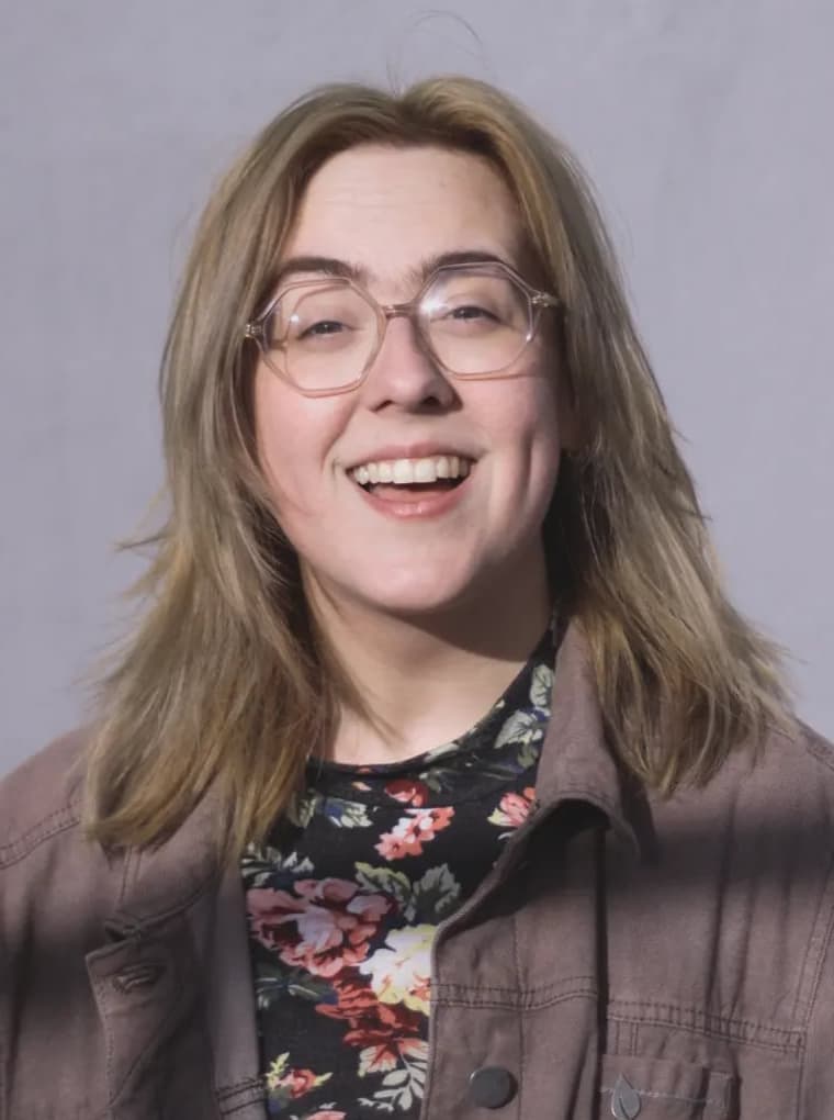 A person with shoulder-length light brown hair and glasses smiles at the camera. They wear a floral top under a brown jacket, standing against a plain light background.