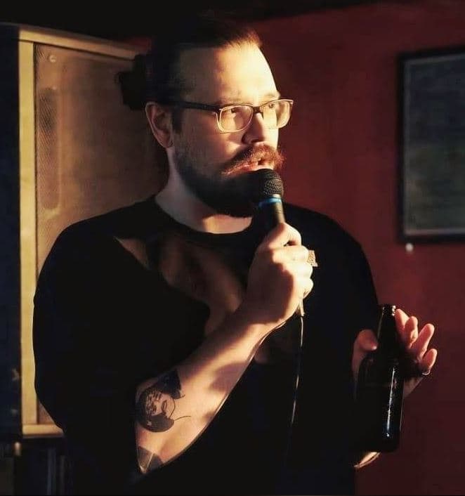 A person with glasses and a beard holds a microphone while speaking on stage, also holding a bottle in the other hand. They wear a dark shirt, and a warm light casts shadows against a wall behind them.