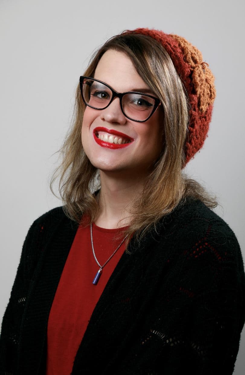 Person wearing black cat-eye glasses and a knitted red-and-brown beanie smiles toward the camera. They wear a red shirt, black cardigan, and a pendant necklace. Studio portrait against a plain light gray background.