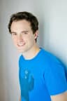 A person with short dark hair smiles at the camera while standing sideways against a light-colored wall. They wear a bright blue T-shirt with a dark graphic on the front. The background is softly blurred, and the portrait is evenly lit with natural-looking light.