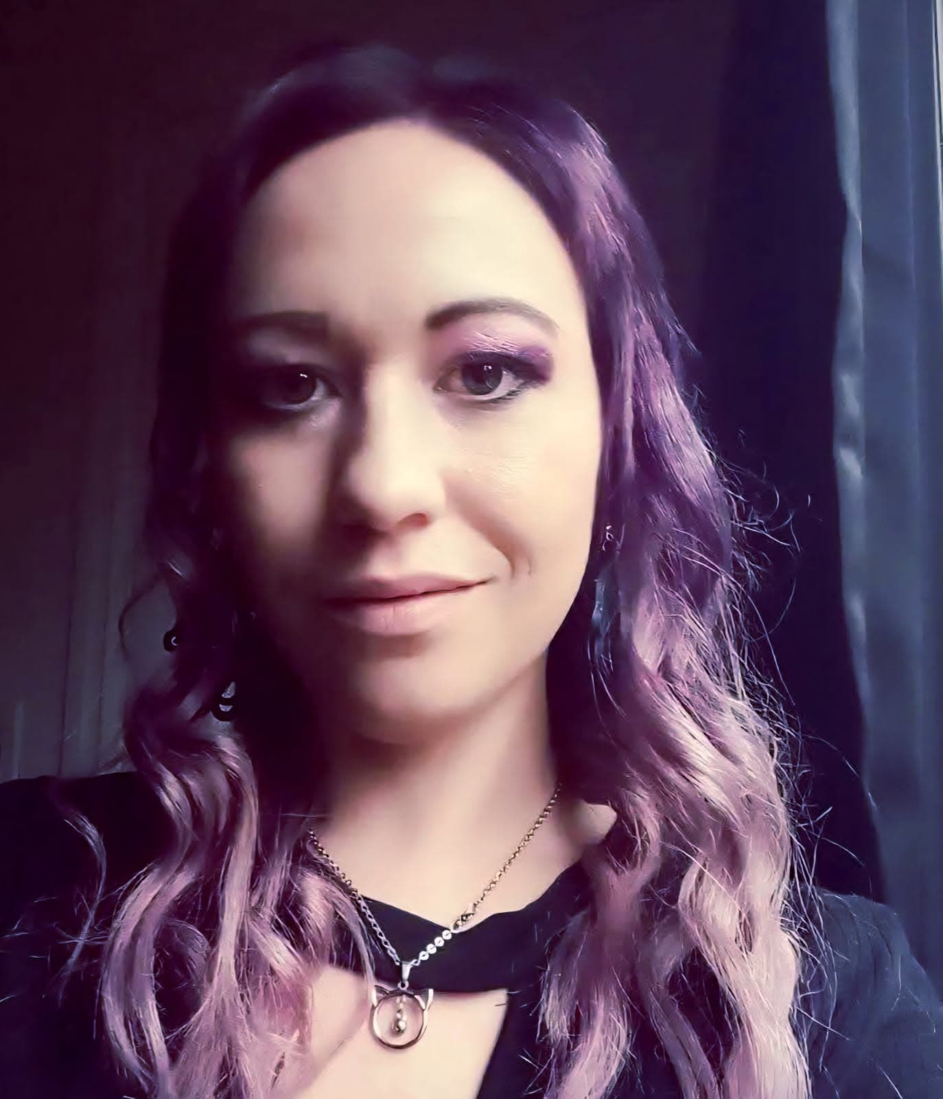 Close-up portrait of a person with long, wavy purple hair, wearing a black top and a necklace with a circular pendant. They face the camera with a neutral expression, with a dark curtain and dim indoor background.
