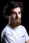 "Studio portrait of a person with dark curly hair and a full beard, wearing a white T-shirt with a small 'Pathshop' logo. The person faces slightly sideways while looking toward the camera against a black background."