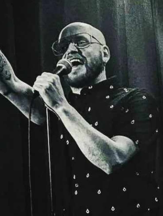 A person with a shaved head and beard performs on stage, holding a microphone and raising one arm. They wear glasses and a short-sleeve patterned shirt. The image is in black and white with a dark curtain in the background.
