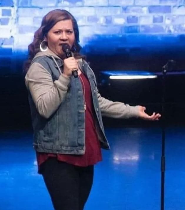 A woman stands on a stage holding a microphone, speaking with one hand extended. She wears a denim vest over a hoodie and a long top, with large hoop earrings. A brick wall and stage lighting are visible in the background.