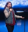 A woman stands on a stage holding a microphone, speaking with one hand extended. She wears a denim vest over a hoodie and a long top, with large hoop earrings. A brick wall and stage lighting are visible in the background.