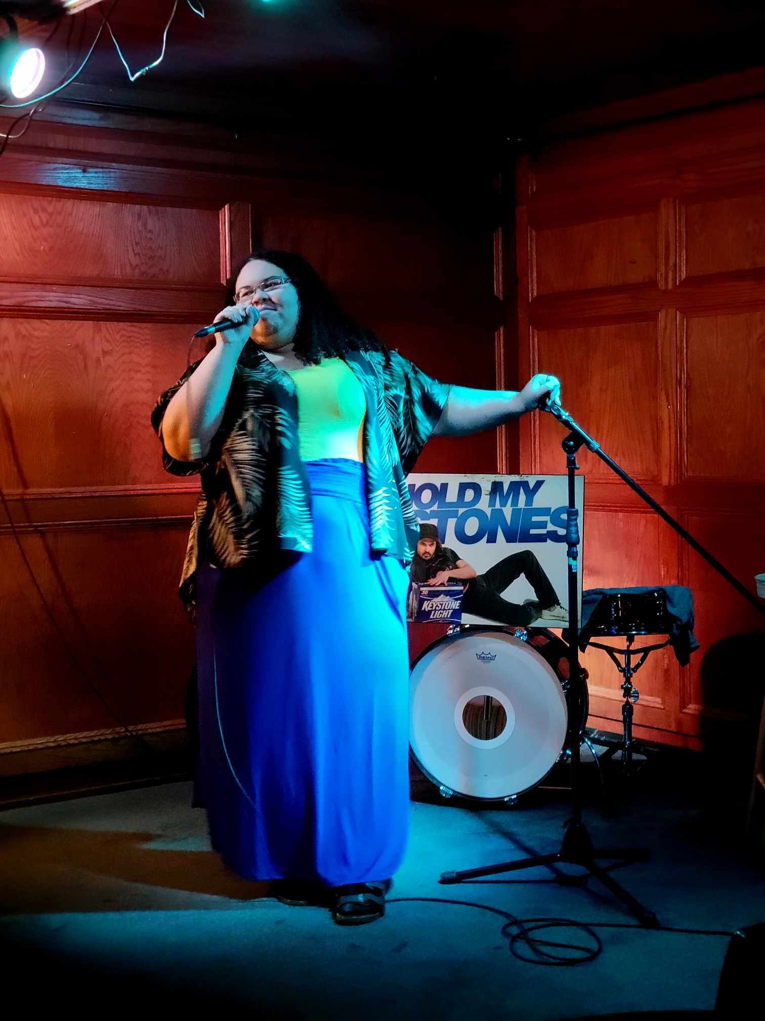 A person performs on a small stage, holding a microphone and standing beside a mic stand. They wear a long blue skirt, a light top, and a patterned shirt. Colored stage lights cast blue and red tones. A drum and a poster reading “HOLD MY STONES” are visible behind them.