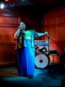 A person performs on a small stage, holding a microphone and standing beside a mic stand. They wear a long blue skirt, a light top, and a patterned shirt. Colored stage lights cast blue and red tones. A drum and a poster reading “HOLD MY STONES” are visible behind them.