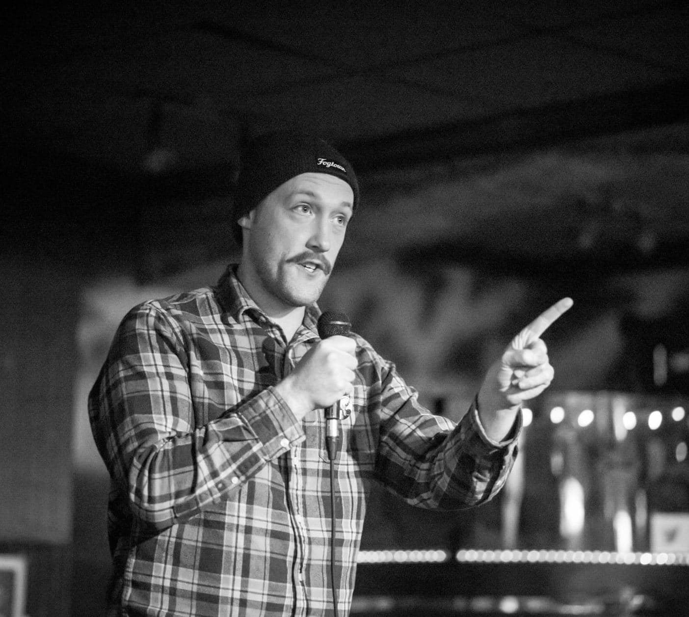 A man wearing a beanie and plaid shirt performs on stage, holding a microphone and pointing to the side. The image is in black and white, with blurred lights and a dim venue in the background.