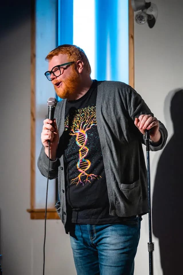 A man with red hair and a beard performs stand-up comedy, holding a microphone and leaning on a mic stand. He wears glasses, a gray cardigan, jeans, and a black T-shirt with a colorful DNA tree design. A lit wall and window are behind him.