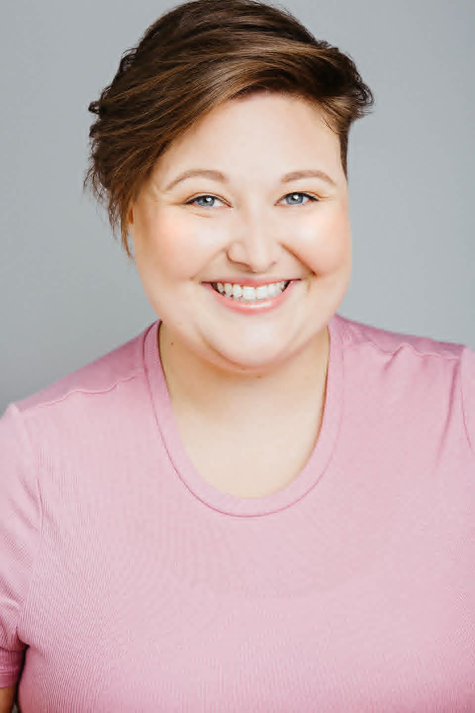 Headshot of a person with short, side-swept brown hair smiling at the camera. They wear a light pink shirt and are positioned against a plain light gray background.