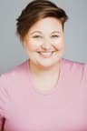Headshot of a person with short, side-swept brown hair smiling at the camera. They wear a light pink shirt and are positioned against a plain light gray background.