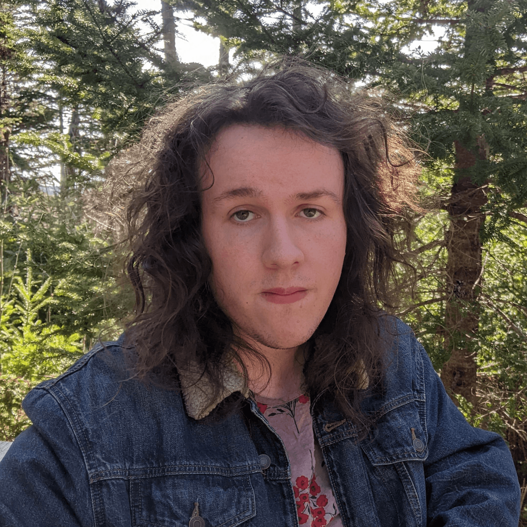 Person with long, wavy dark hair sits outdoors among evergreen trees, facing the camera. They wear a blue denim jacket with a light-colored collar over a patterned shirt. Sunlight filters through the trees behind them.  Extended Description: A close-up outdoor portrait of a person seated in a wooded area. They have shoulder-length, wavy dark hair and are looking directly at the camera. They are wearing a blue denim jacket with a light fleece-style collar and a shirt with a red floral pattern visible at the neckline. Evergreen branches and tree trunks fill the background, with bright sunlight shining through the foliage and creating highlights around the hair and trees. No visible text appears in the image.