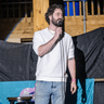 A man with curly hair and a beard stands on a small stage holding a microphone, wearing a light sweater and jeans. A stool sits beside him. Behind him are wooden railings and a colorful banner, suggesting a live comedy performance.