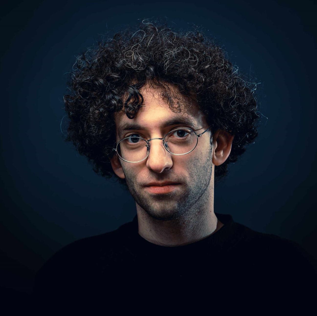 A person with short, curly dark hair and round glasses looks directly at the camera against a dark blue background. They wear a black top and have a neutral expression.