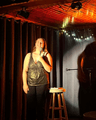 A smiling woman stands on a small stage holding a microphone beside a stool with papers, framed by dark curtains and warm string lights.