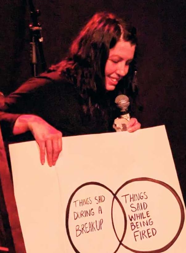 A person on stage holds a microphone and a large poster with a Venn diagram. The circles read “Things said during a breakup” and “Things said while being fired.” Red stage lighting and a dark curtain are in the background.