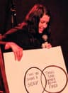A person on stage holds a microphone and a large poster with a Venn diagram. The circles read “Things said during a breakup” and “Things said while being fired.” Red stage lighting and a dark curtain are in the background.