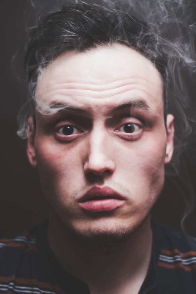 Close-up portrait of a person with dark hair, facing forward. Wisps of smoke surround their head and forehead. Their eyes are wide open, lips slightly parted. The background is dark, and they wear a striped shirt.