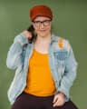 A person wearing glasses and a red cap looks slightly to the side while holding their hair. They wear a light blue distressed denim jacket over a mustard-yellow top, posed against a plain green background.