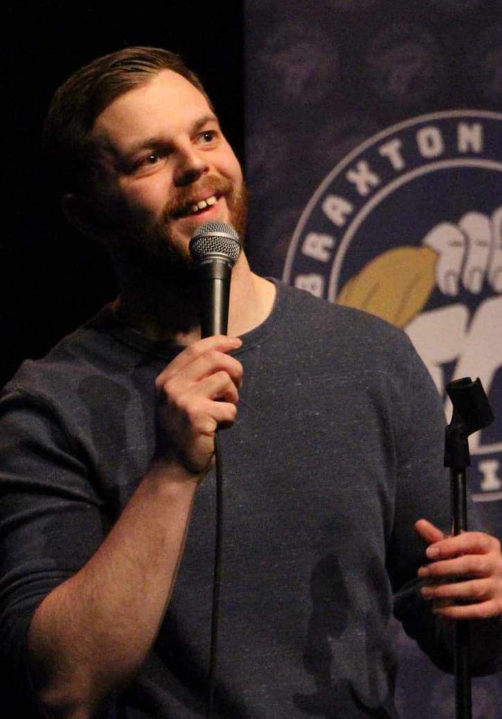 A man with short hair and a beard smiles while holding a microphone on stage. He wears a dark long-sleeve shirt and stands near a mic stand, with a partially visible logo backdrop behind him.