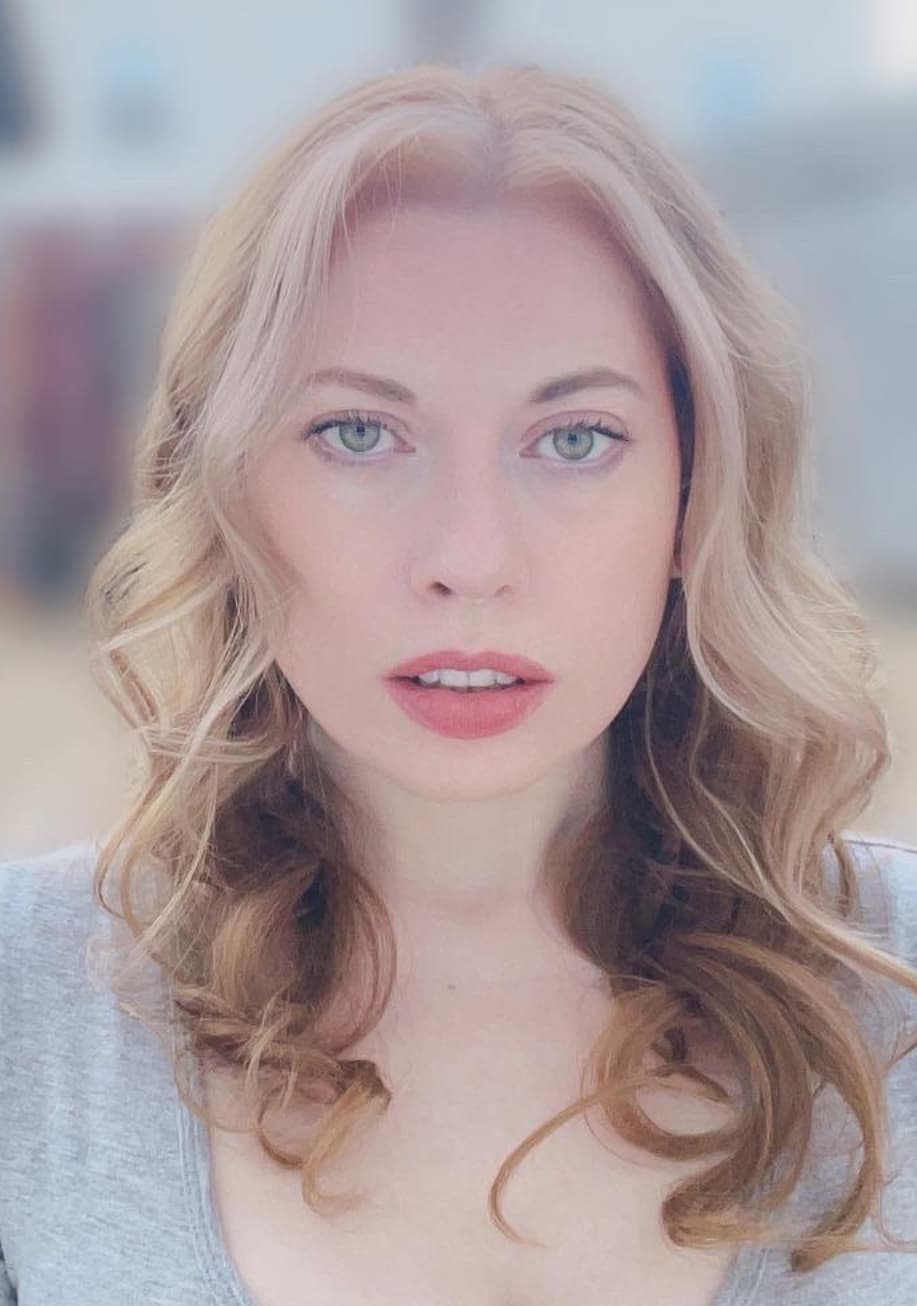 A close-up portrait of a person with shoulder-length wavy blonde hair looking directly at the camera. They have light-colored eyes and wear a light gray top, with a softly blurred background.