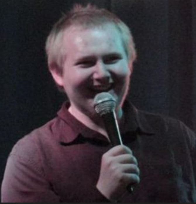 A person stands on stage holding a microphone and smiling while speaking. They wear a dark button-up shirt. The image is slightly grainy with a dark curtain background and colored stage lighting.